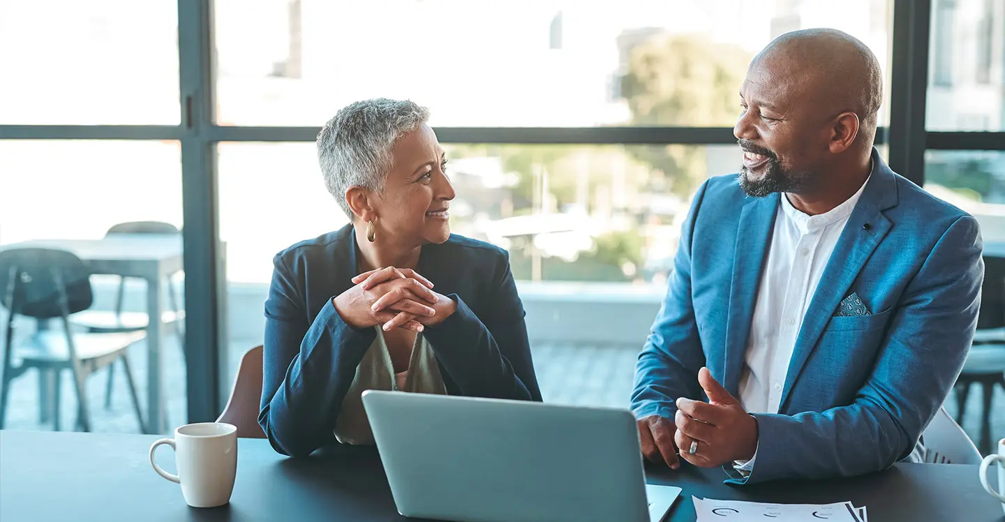 Man and senior woman having a discussion in windowed office over laptop computer.
